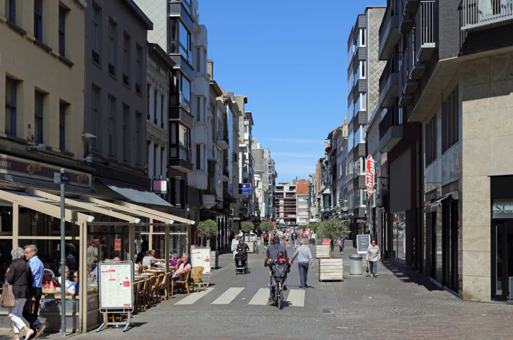 Shoppers and a cyclist on the car-free Adolf Buylstraat in Ostend on a sunny day, lined with shops and a cafe terrace