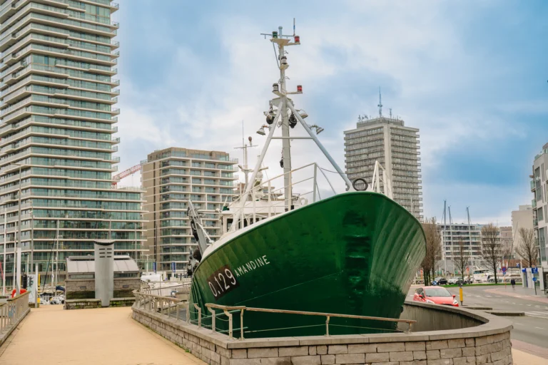 The green-hulled trawler Amandine O.129 in its dry dock on the quayside, with apartment towers behind