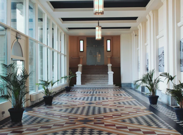 Glazed Art Deco gallery with a patterned tiled floor, potted palms and a staircase leading up to the hotel