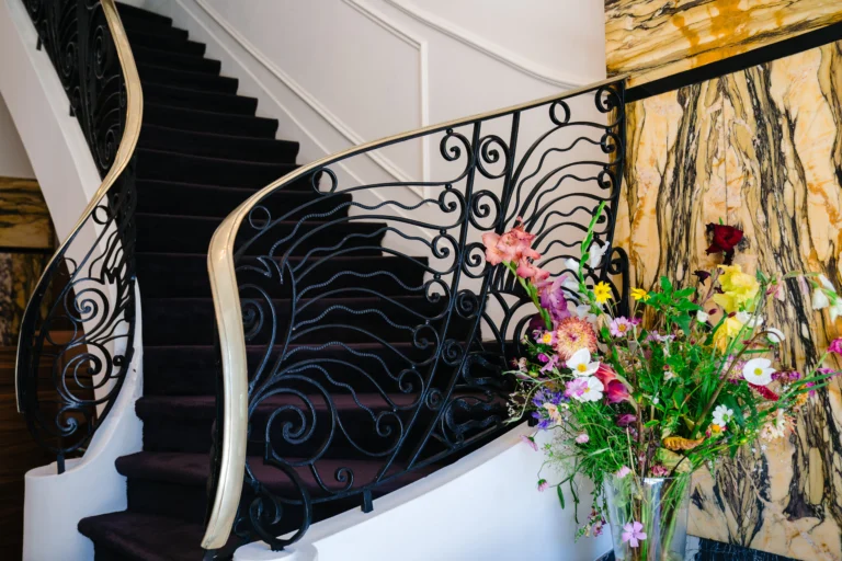 Curving Art Deco staircase with a wrought-iron balustrade, marble wall and a vase of fresh flowers