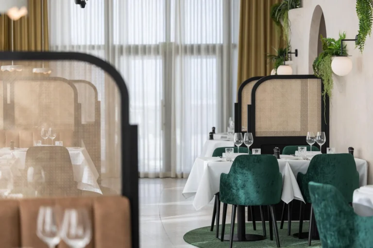 Dining room with cane-backed screens, tables laid with white cloths and green velvet chairs in front of tall curtained windows