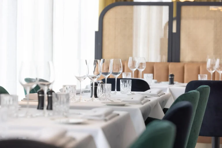 Rows of tables laid with white cloths, glasses and folded napkins, with green and navy chairs and a cane-backed banquette behind