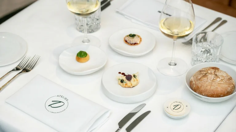 Table set at Atelier D with white linen, a napkin printed with the Atelier D logo, two glasses of white wine, small bites on white plates and a loaf of bread
