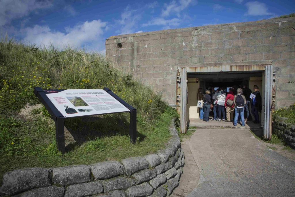 Visitors gathered at the open steel doors of a concrete bunker set into a grassy dune, beside an information panel