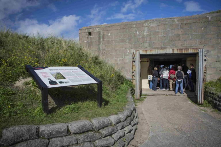 Visitors gathered at the open steel doors of a concrete bunker set into a grassy dune, beside an information panel