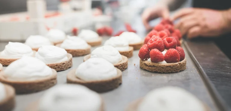 Small tartlets topped with cream and fresh raspberries on a metal tray, being finished by hand