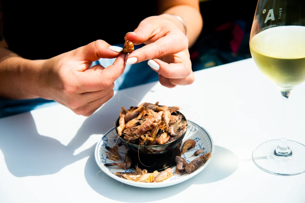Hands peeling a small shrimp from a bowl of shrimps on a sunlit white table, with a glass of white wine alongside