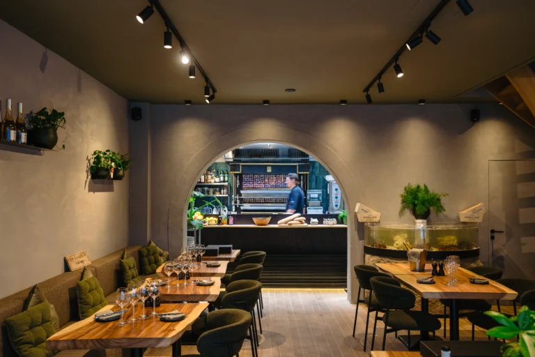 The dining room at Bar Foubert, with wooden tables and green chairs under track spotlights, leading to an arched opening onto the open kitchen