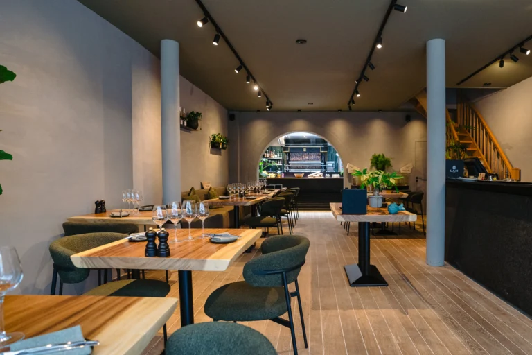 Looking down the long dining room at Bar Foubert, with wooden tables, green chairs, a wooden staircase on the right and the open kitchen at the back