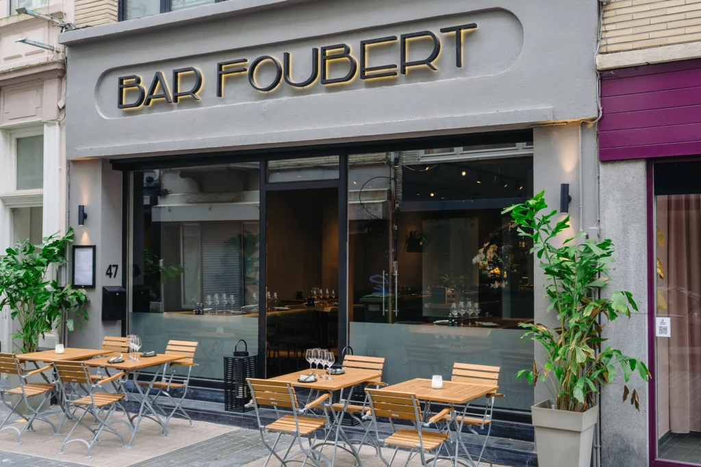 The grey frontage of Bar Foubert on Christinastraat with gold lettering above large windows, and wooden bistro tables and chairs set out on the pavement