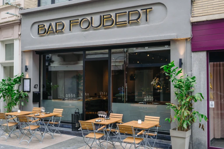 The grey frontage of Bar Foubert on Christinastraat with gold lettering above large windows, and wooden bistro tables and chairs set out on the pavement