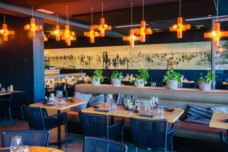 Dining room with wooden tables, banquette seating, orange cross-shaped pendant lights and a long black-and-white beach scene along the wall