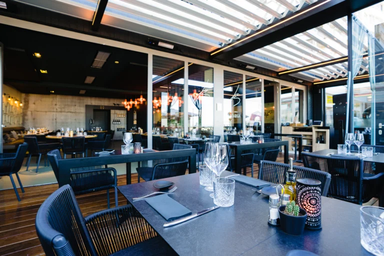 Dining area under a slatted retractable roof with glass screens, dark tables laid with glasses and cutlery and a bar in the background