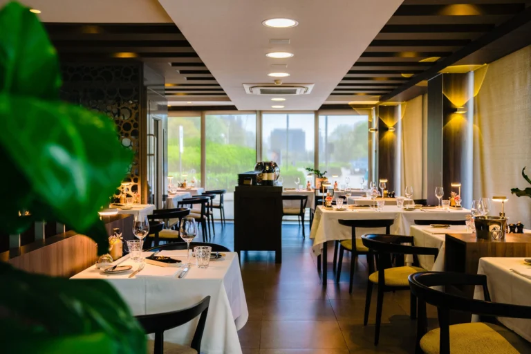 Dining room facing the tall windows, with white-clothed tables laid for service and a large green plant in the foreground