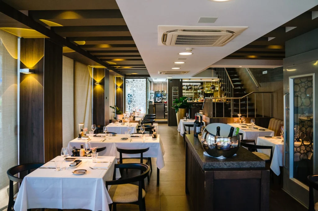 Long dining room at Bistro Mathilda with white-clothed tables, black chairs and wall lights, and a bar and staircase at the far end