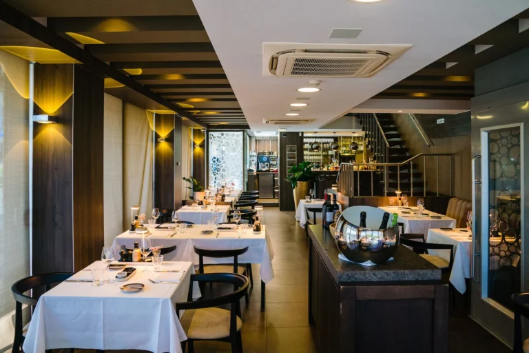 Long dining room at Bistro Mathilda with white-clothed tables, black chairs and wall lights, and a bar and staircase at the far end