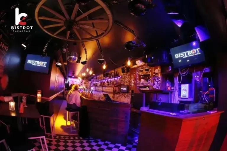 Dimly lit bar interior with a checkerboard floor, a wagon-wheel light fitting and a long bar glowing in purple and orange light