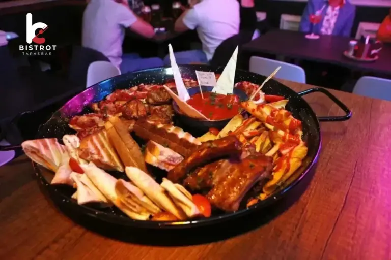 Large black pan of mixed tapas with ribs, toasted wraps, cured ham and a pot of tomato dip on a wooden table