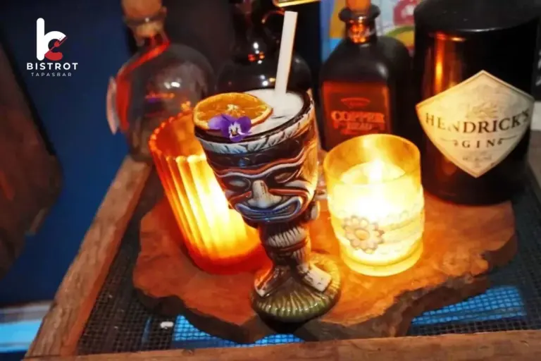 A tiki-mug cocktail garnished with dried orange and a violet flower between two candles on a wooden bar, with gin and rum bottles behind