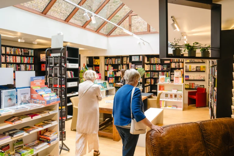 Bookshop interior under a sloping glass roof, with tall bookcases, display tables and two customers browsing