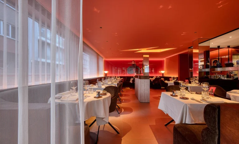 View across the dining room through sheer curtains, with white-clothed tables under a red-lit ceiling