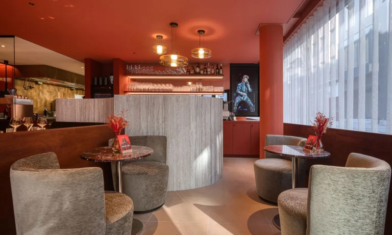 Lounge corner with grey armchairs and round tables in front of a curved bar and lit shelves
