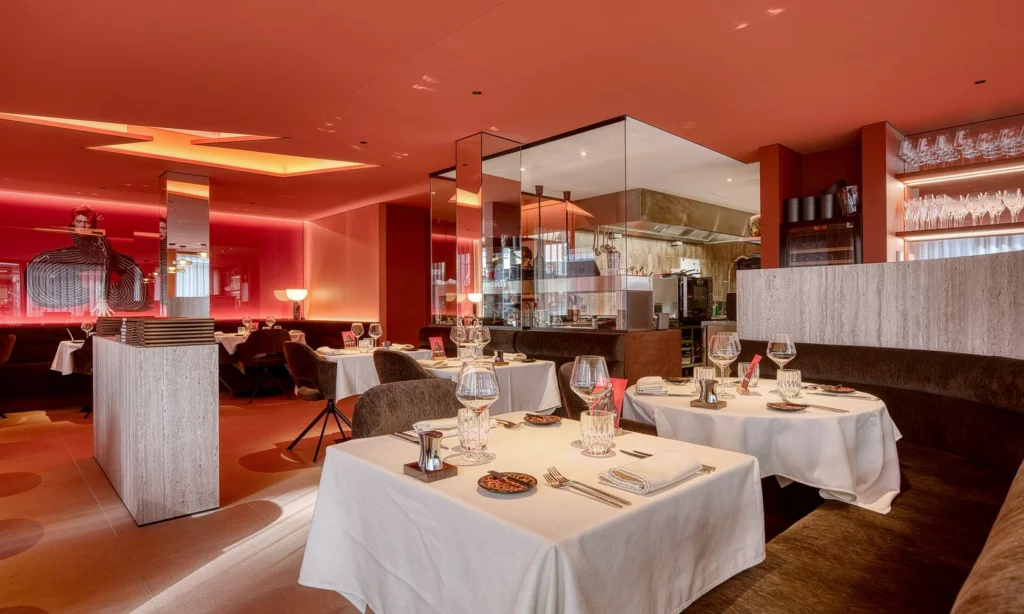 Dining room lit in warm red, with white-clothed tables, dark banquettes and shelves of glasses behind glass partitions