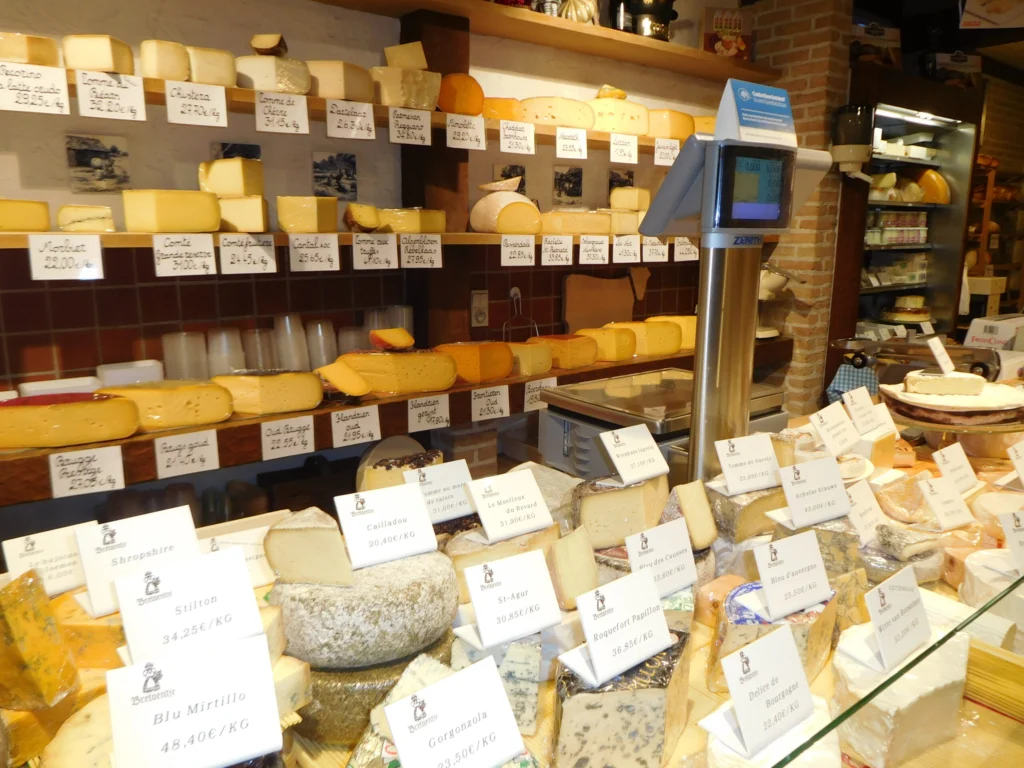 Glass cheese counter at Bretoentje full of labelled wedges and wheels, with shelves of hard cheeses and a weighing scale behind