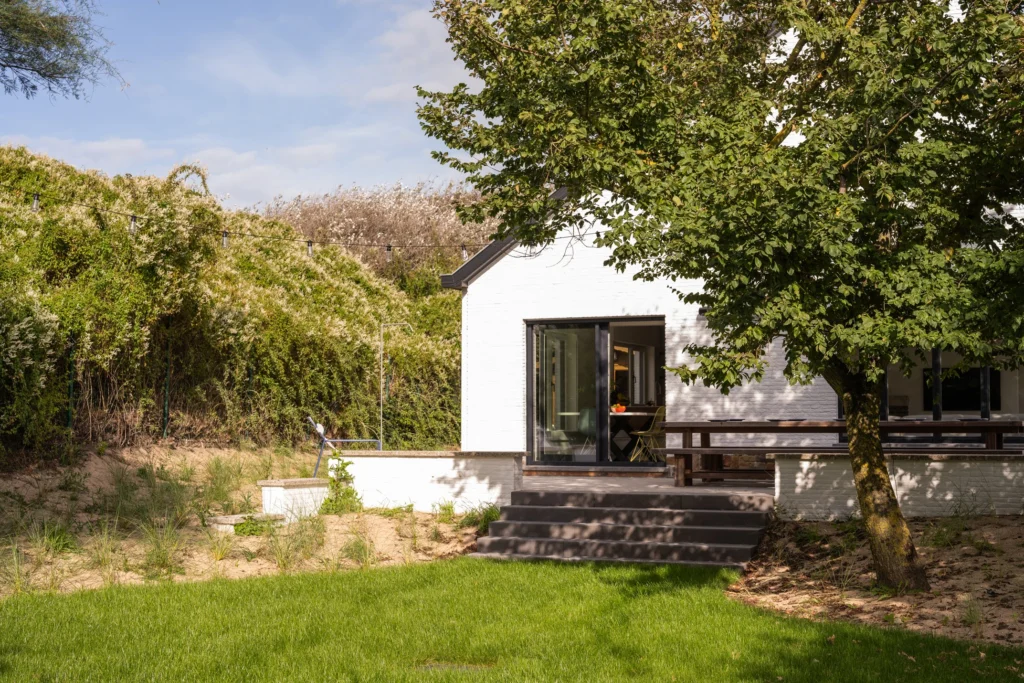 White-painted holiday house with a terrace and lawn, framed by dune vegetation and a large tree