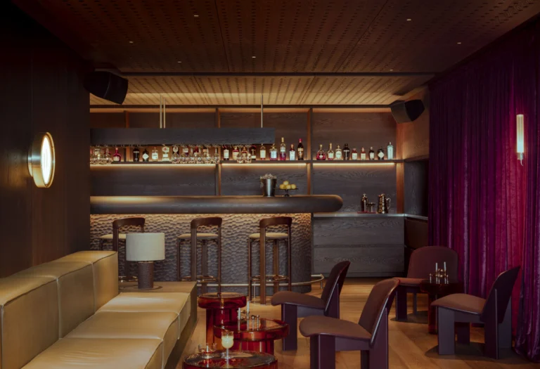 Curved bar counter with high stools in front of a backlit shelf of spirits, with a leather banquette and burgundy chairs in the foreground