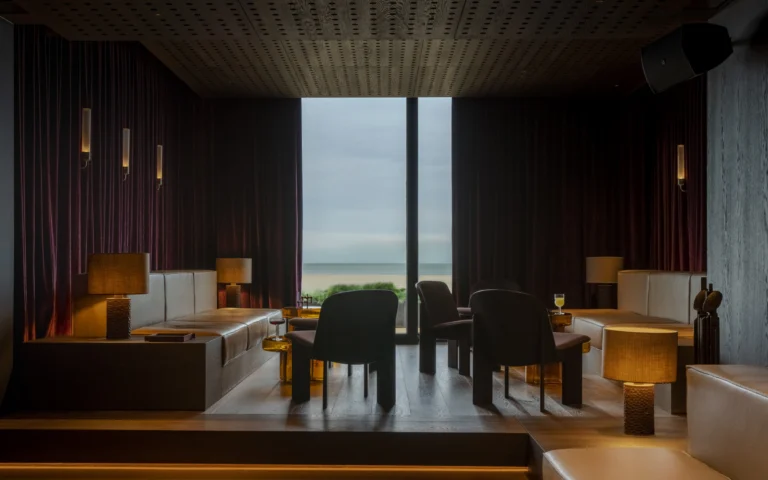 Dimly lit lounge at C-Bar with leather banquettes, table lamps and red velvet curtains around a tall window looking out to the beach and the sea
