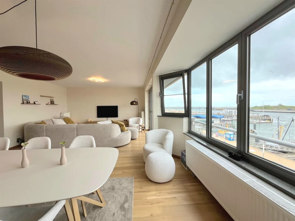 Living area with a curved grey sofa and white armchairs beside corner windows overlooking the harbour and marina