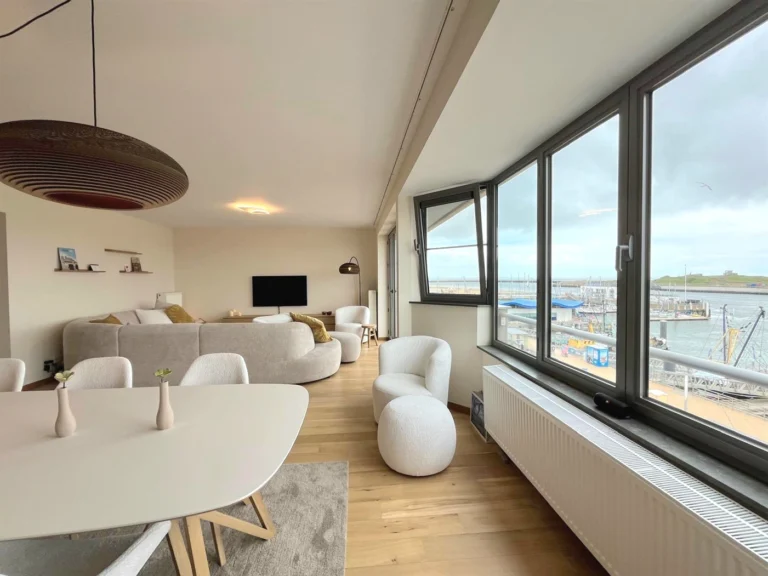 Living area with a curved grey sofa and white armchairs beside corner windows overlooking the harbour and marina
