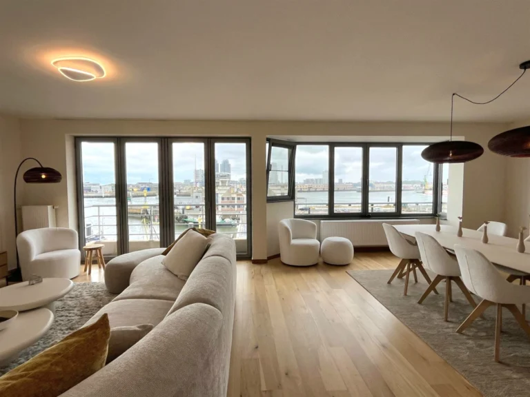 Open-plan lounge and dining room with a wide sofa, oval table and a long run of windows onto the harbour
