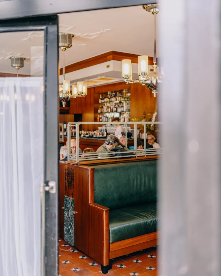 Looking through an open glass door into Café Du Parc, with a green leather banquette, wood-panelled bar and chandeliers
