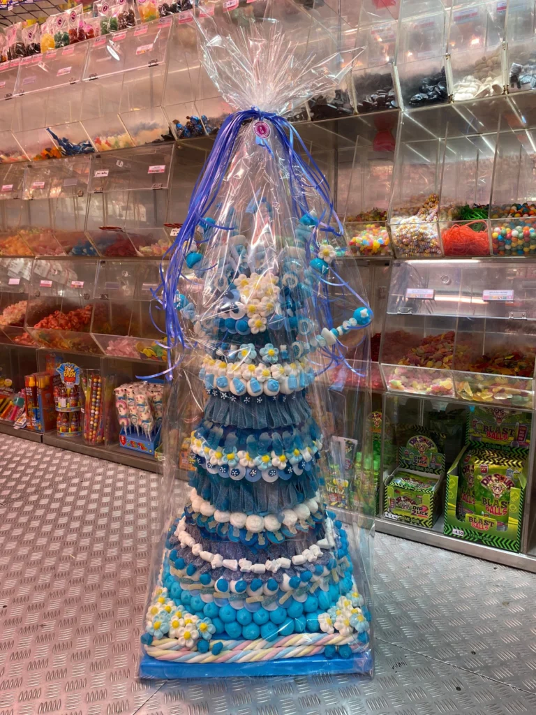 A tall tiered candy tower wrapped in clear cellophane with a purple ribbon, built from blue, white and yellow sweets and marshmallows, standing on the shop floor in front of walls of clear dispensers full of sweets
