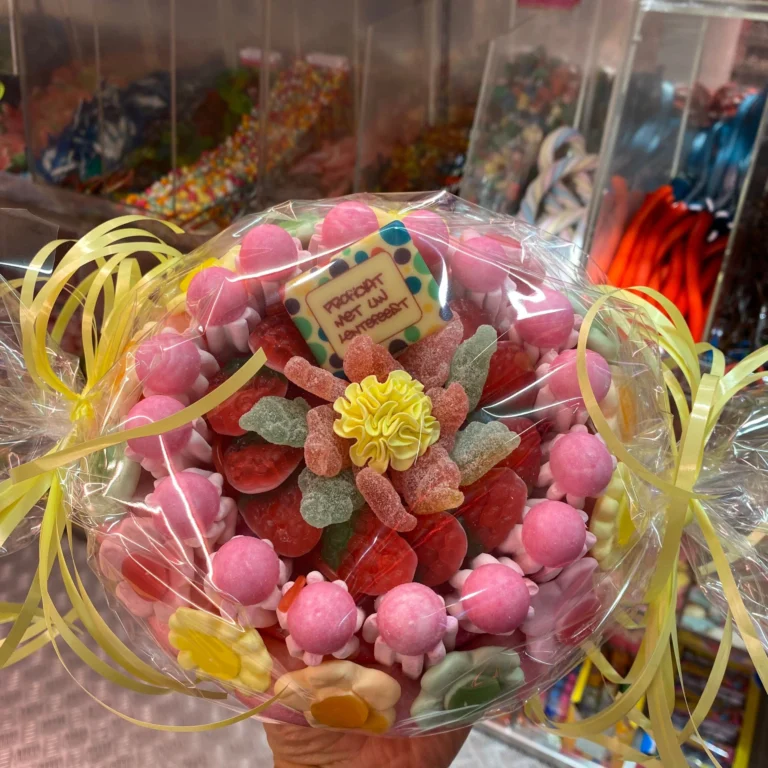 A round candy bouquet wrapped in cellophane with yellow ribbon curls, topped with pink gummies, sour sweets and a yellow sugar flower, held up in front of shelves of pick-and-mix sweets