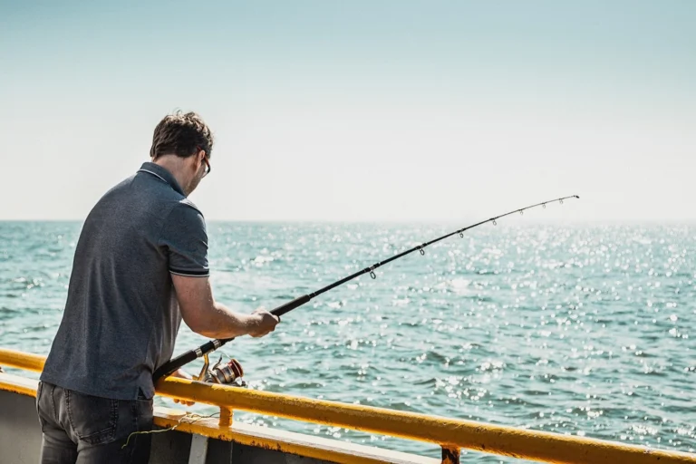 Man fishing with a long rod over the yellow railing, sunlight glittering on the sea