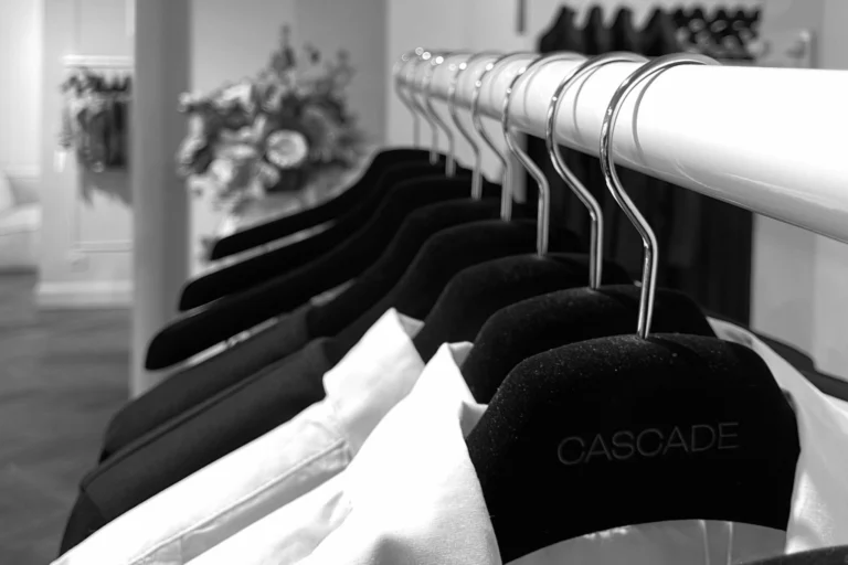 Black-and-white close-up of black velvet hangers with chrome hooks on a white rail, one embossed with the Cascade name, with a vase of flowers blurred behind