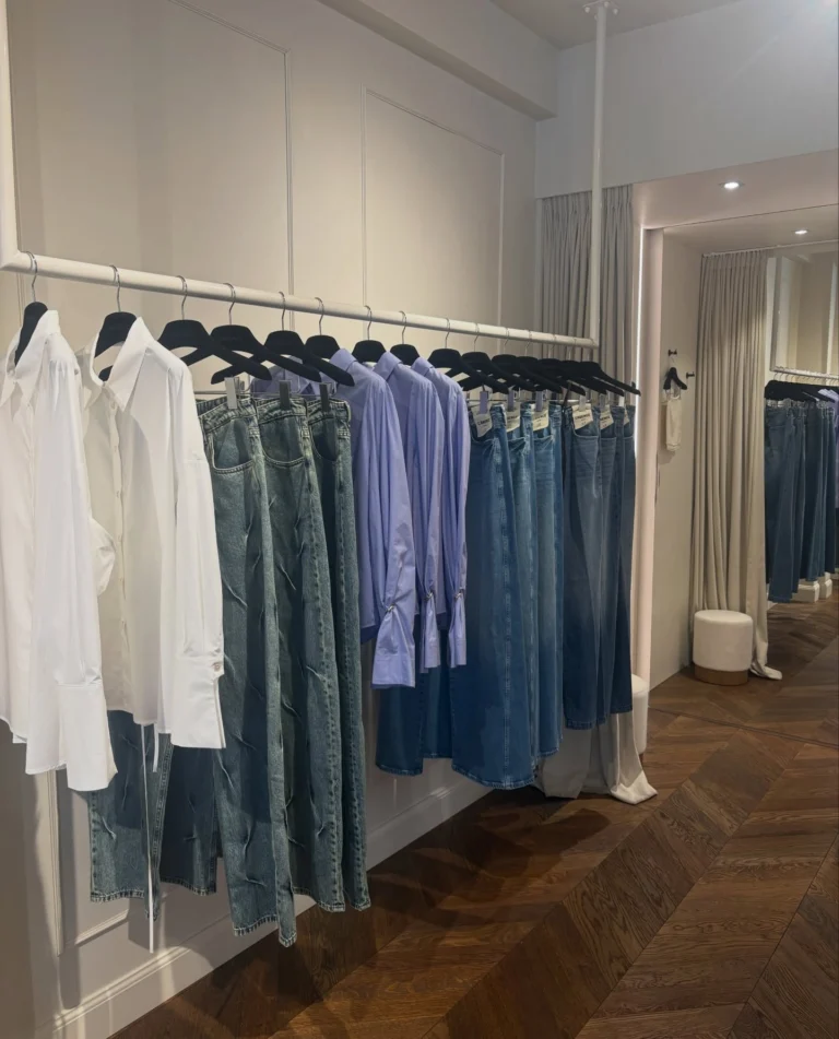 White shirts, lilac blouses and a row of blue jeans hanging on a white rail in a panelled room with a herringbone wooden floor and curtained fitting room