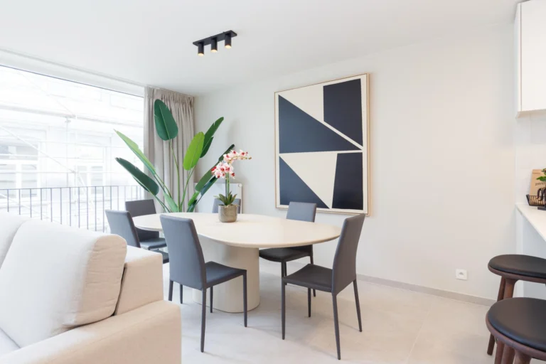 Round dining table with four grey chairs beside a large window, a leafy plant and black-and-white artwork