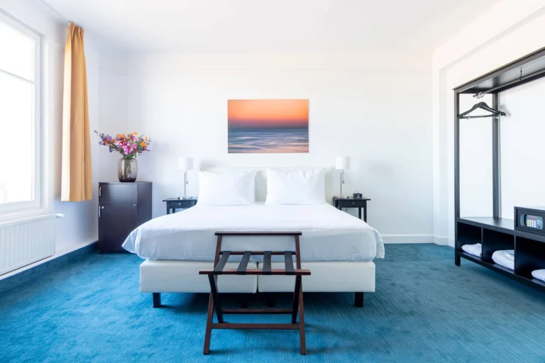 Bright double room with a blue carpet, white bed, open wardrobe rail and a sunset seascape above the bed