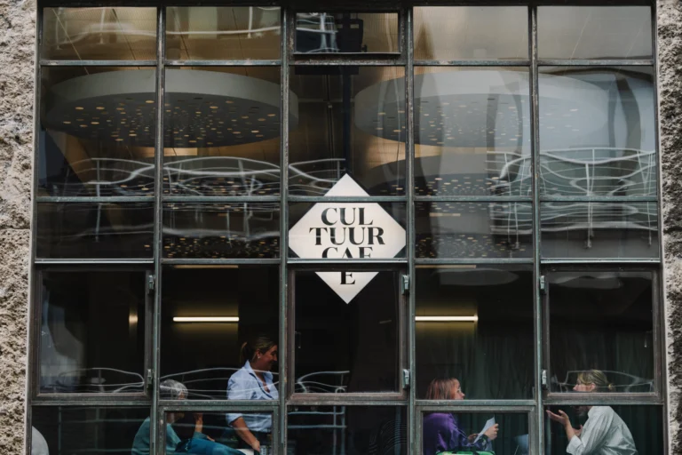 The glazed steel-framed front of the café with a diamond-shaped CultuurCafé sign and guests seated inside