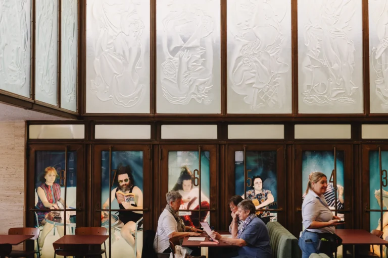 Guests at wooden tables under tall panels of relief-patterned glass in the café