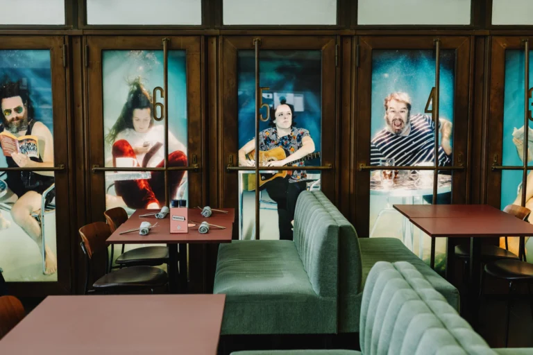 The dining room with green banquette seating and red-brown tables in front of tall glazed doors dressed with large portrait panels