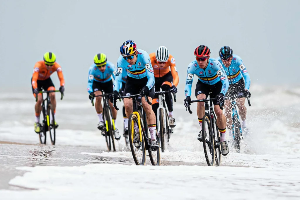Cyclo-cross riders splashing through shallow surf on a grey North Sea beach during a championship race