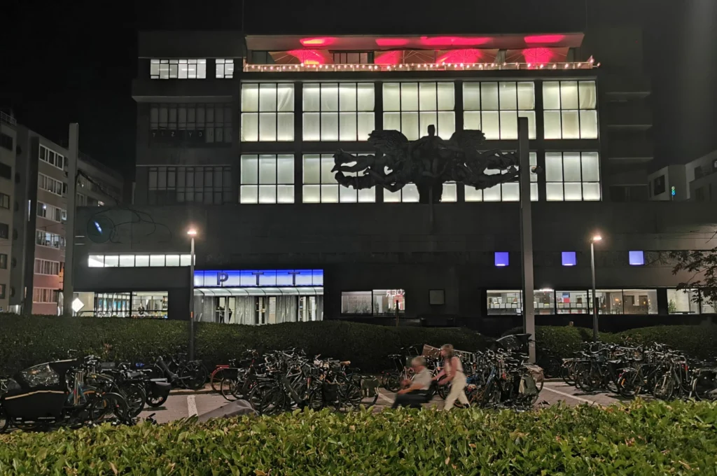 Modernist façade of De Grote Post lit up at night, with large glass windows, Cantré's bronze sculpture and parked bicycles in front