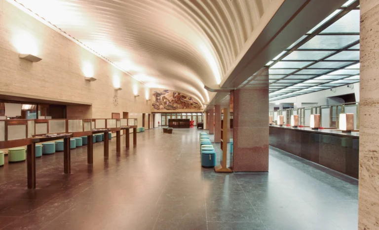 Long former post office counter hall with a curved ceiling, stone and wooden counters and a mural at the far end