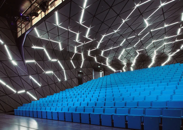 Empty theatre hall with rows of blue seats under a dark faceted ceiling crossed by zigzag light strips
