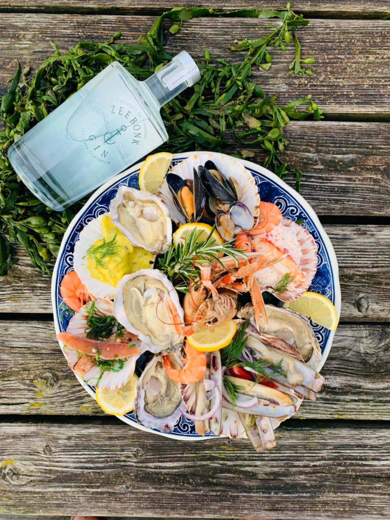 A blue-and-white plate piled with oysters, mussels, razor clams, langoustines and prawns with lemon wedges, on a weathered wooden table beside a bottle of gin and fresh seaweed, seen from above